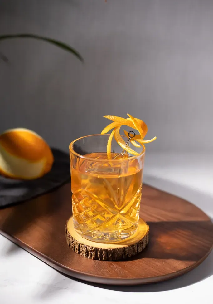 un cóctel dorado en un vaso bajo con una elegante piel de naranja.' title=
