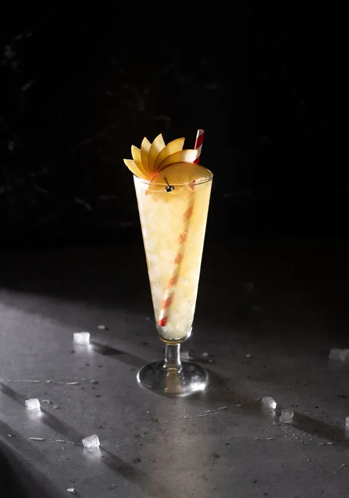 un vaso pilsner lleno de hielo picado y líquido dorado con una pajita de papel.' title=