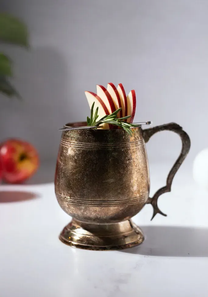 primer plano de una taza de mula deslustrada con un abanico de manzana y una ramita de romero.' title=