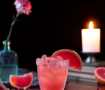 Rosafarbenes Getränk in einem kurzen Glas mit Grapefruitschnitzgarnitur neben einer Kerze.' loading='lazy' title=