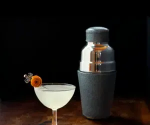 Vintage kupé sklenice naplněná mlhavou kapalinou vedle kůží zabalené koktejlové šejkru.' loading='lazy' title=