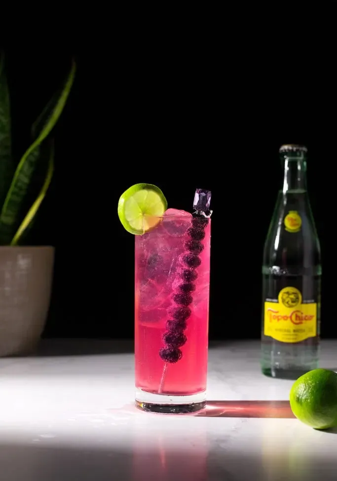 un vaso alto de color rosa brillante junto a una botella de Topo Chico.' title=