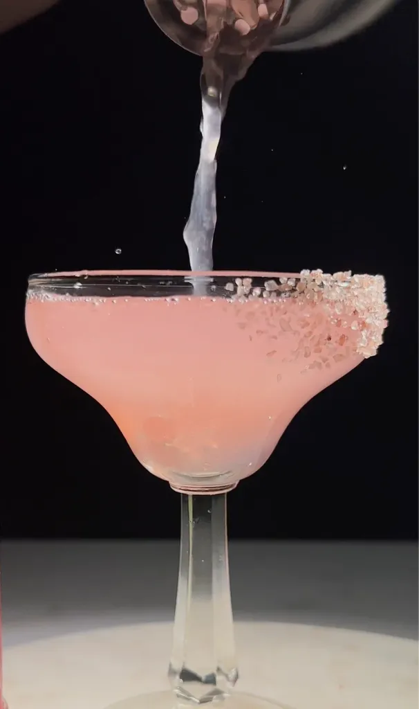 versando il liquido rosa in un bicchiere da margarita bordato di sale rosa.' title=