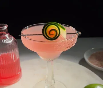 cocktail rosa in un bicchiere di margarita accanto a un lime e una bottiglia di vetro di sciroppo rosa.' title=