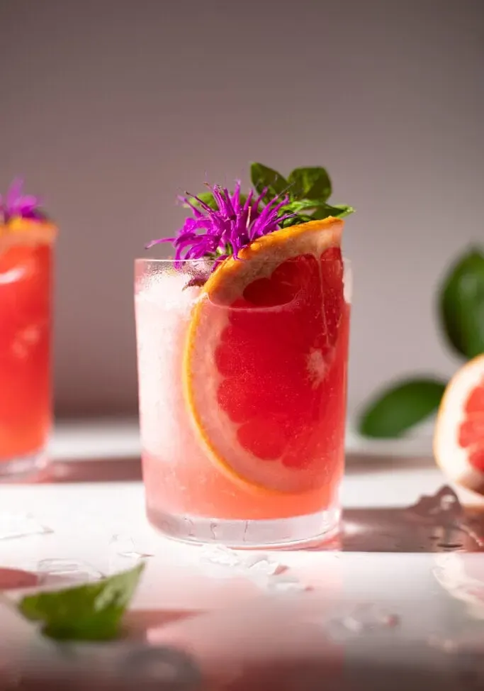 Nahaufnahme eines rosa gefrorenen Getränks mit Grapefruitscheibenblüten und Basilikumblättern.' title=
