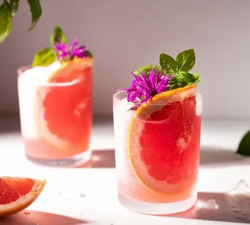zwei Gläser mit Grapefruitscheiben, rosa Blüten und Basilikumblättern.' title=