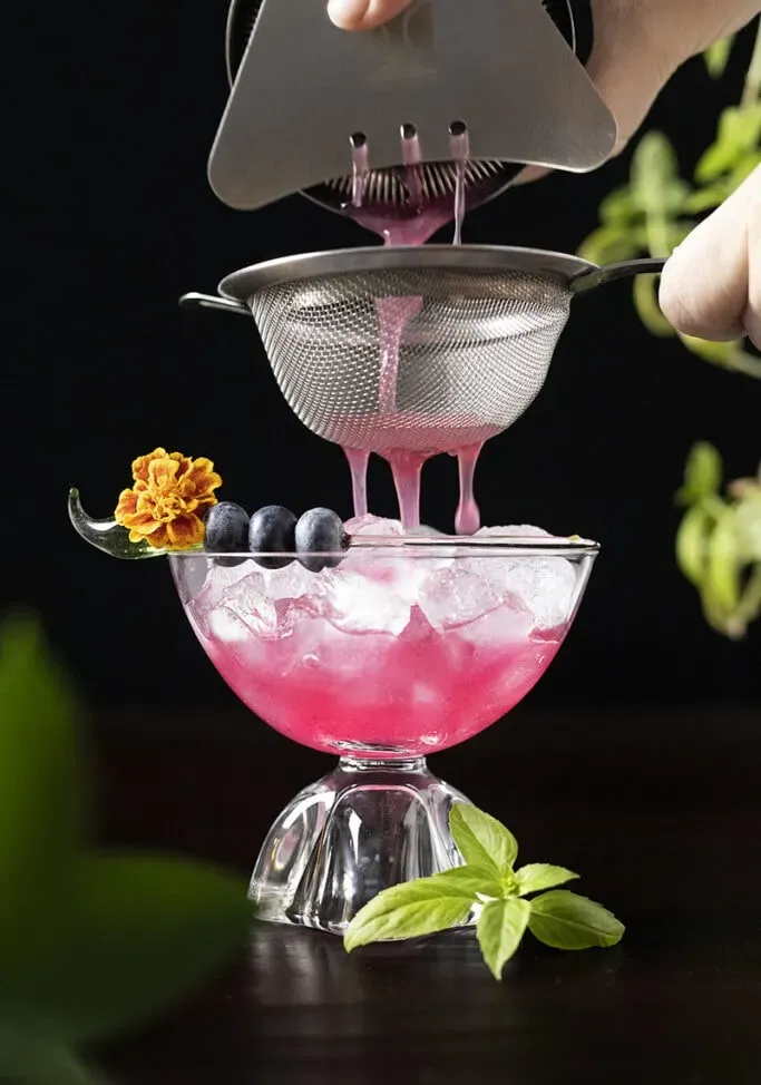 coar uma bebida rosa através de uma peneira de malha fina em um copo.' loading='lazy' title=