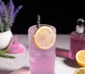 jasnofioletowy napój w szklance typu highball obok cytryn i butelki fioletowego syropu.' title=