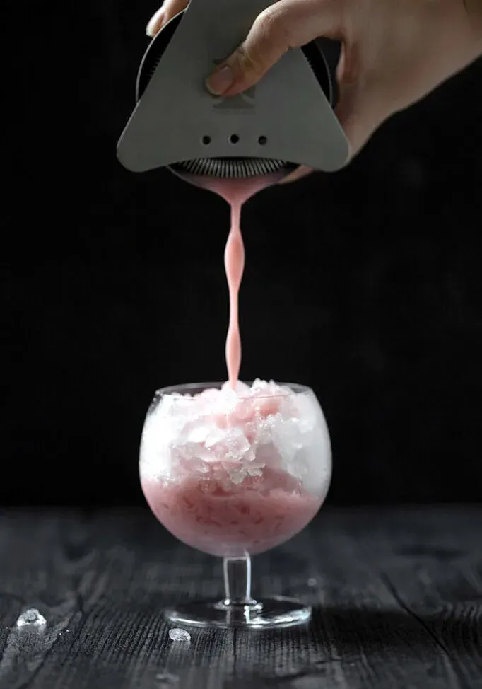 מזיגת נוזל ורוד חלבי משייקר קוקטיילים לכוס מלאה בקרח.' loading='lazy' title=
