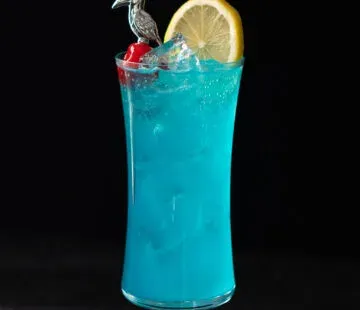 bevanda blu in un bicchiere da uragano con una ruota di limone e un contorno di ciliegie da cocktail.' title=