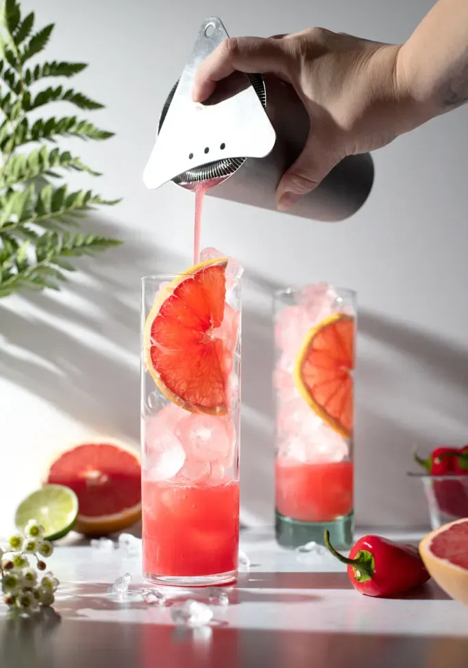 przecedzając różowy płyn do szklanek typu highball wypełnionych lodem.' loading='lazy' title=