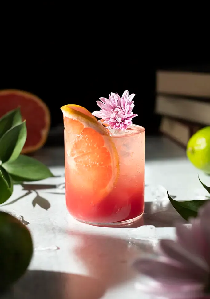 bebida roja en un vaso bajo con una rodaja de pomelo y una flor rosa.' loading='lazy' title=