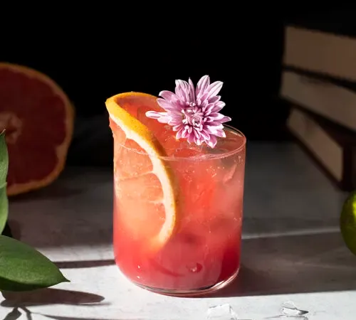 un vaso bajo lleno de líquido rosa y una rueda de pomelo' title=
