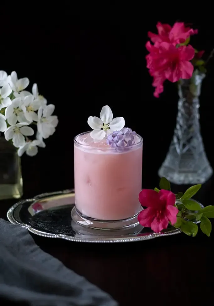 różowy mleczny napój z jadalnymi kwiatami w szklance z kamieniami' title=