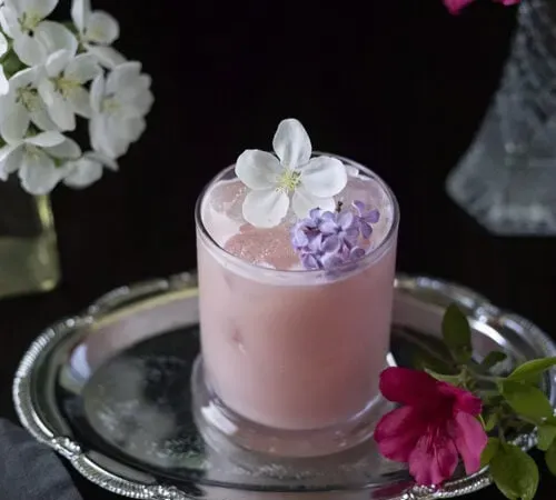 różowy mleczny napój w szklance z kamienia otoczonej kwiatami' title=