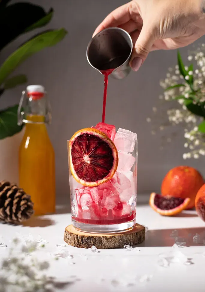 vertiendo jugo de naranja sanguina en un vaso lleno de hielo y una rueda de naranja sanguina.' loading='lazy' title=