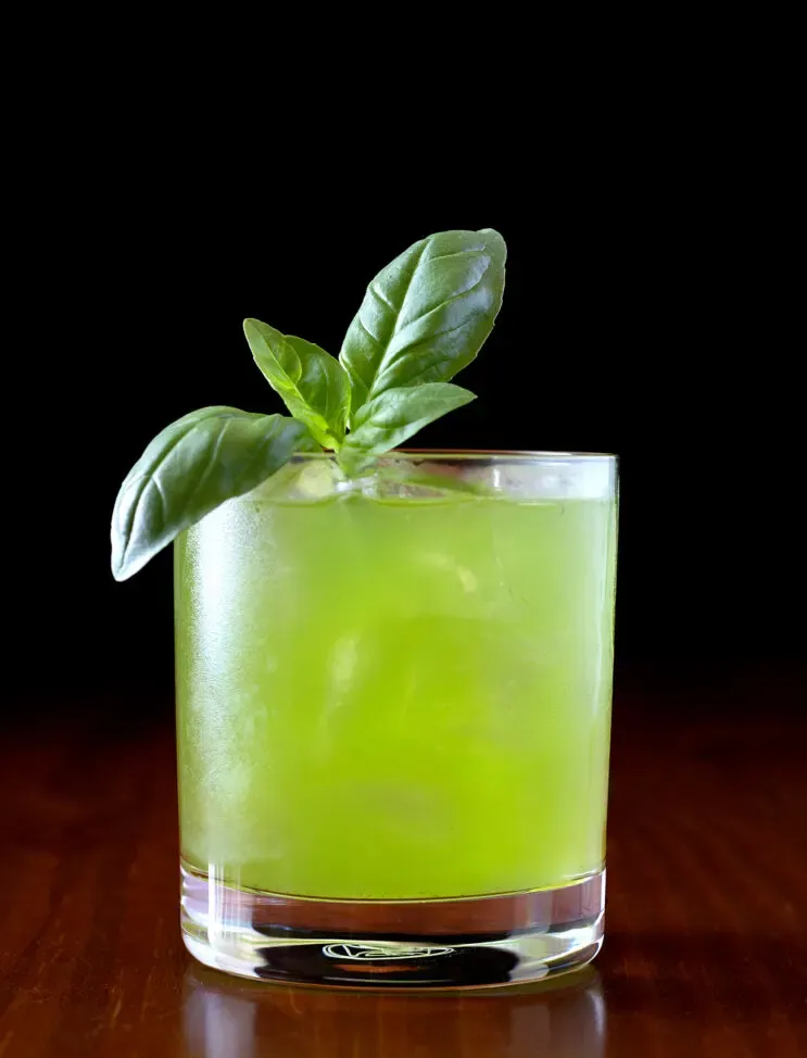 bebida verde en un vaso bajo adornado con una ramita de albahaca.' loading='lazy' title=