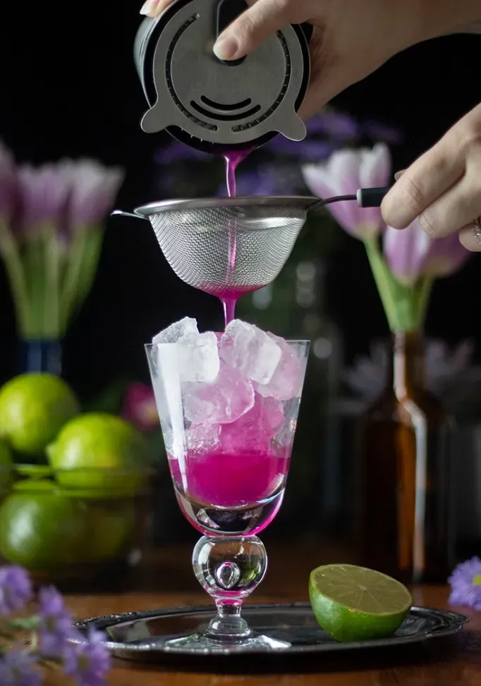 Gießen Sie rosa Flüssigkeit durch ein feinmaschiges Sieb in ein mit Eis gefülltes Glas.' title=