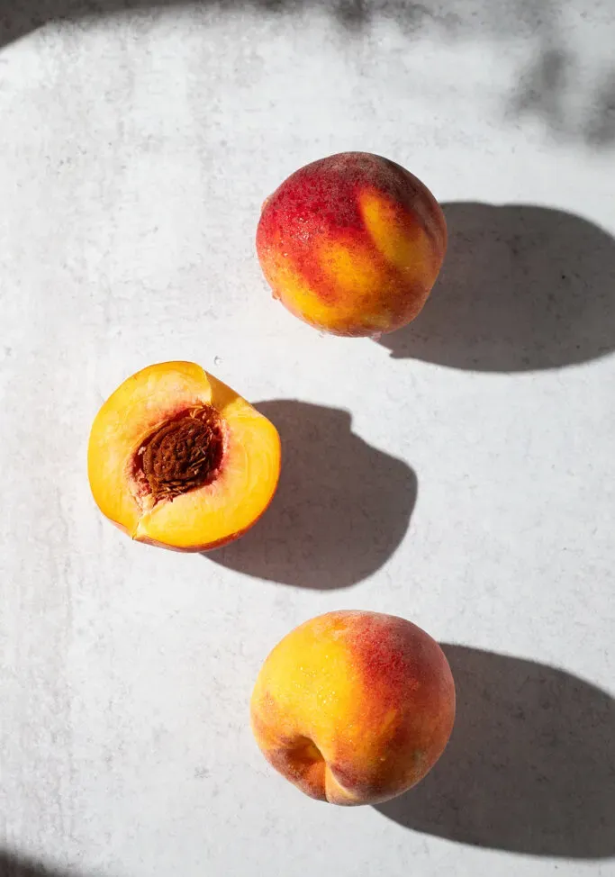 trois pêches jaunes sur une table blanche au soleil.' loading='lazy' title=