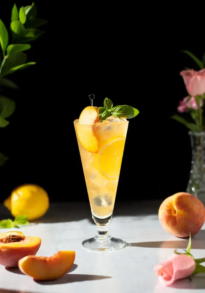 boisson orange avec tranche de pêche à côté de pêches et de roses roses.' title=