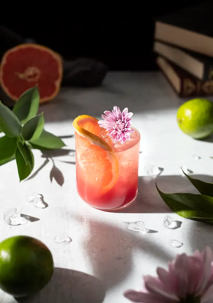 bebida roja con una rodaja de pomelo junto a hojas verdes de lima y una pila de libros.' title=