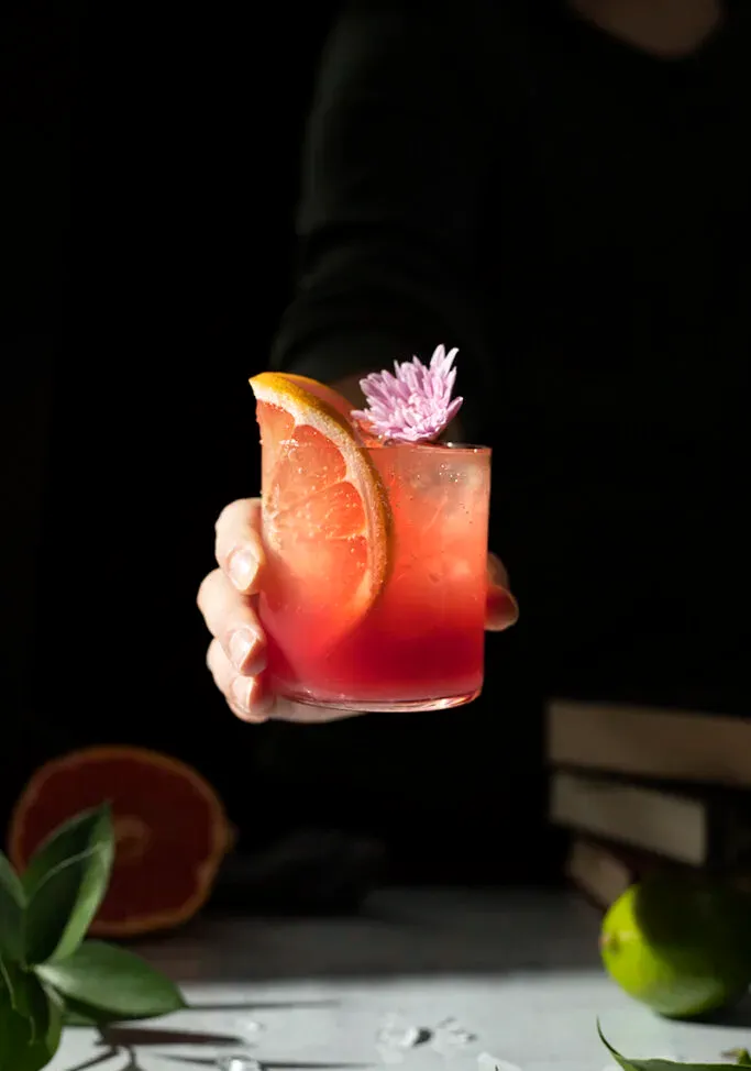 una mano sosteniendo un vaso de líquido rosa bajo el sol.' title=