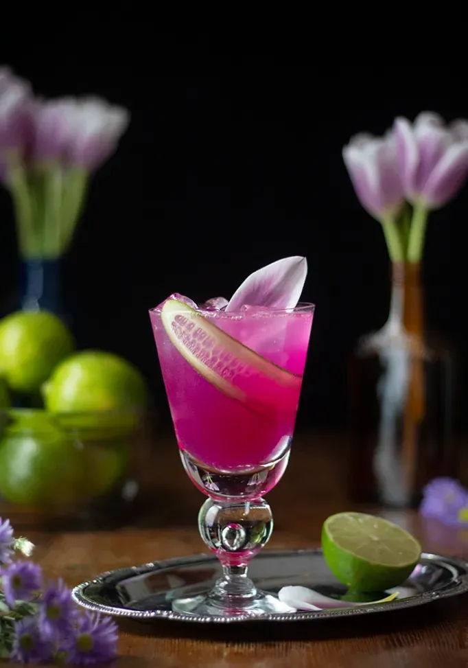 bebida rosa choque em uma bandeja de prata ao lado de uma tigela de limão e flores roxas.' title=