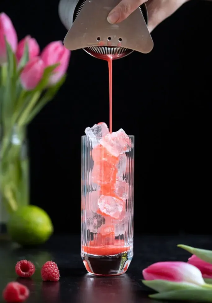 Cocktailshaker, der rote Flüssigkeit in ein mit Eis gefülltes Longdrinkglas abseiht' loading='lazy' title=