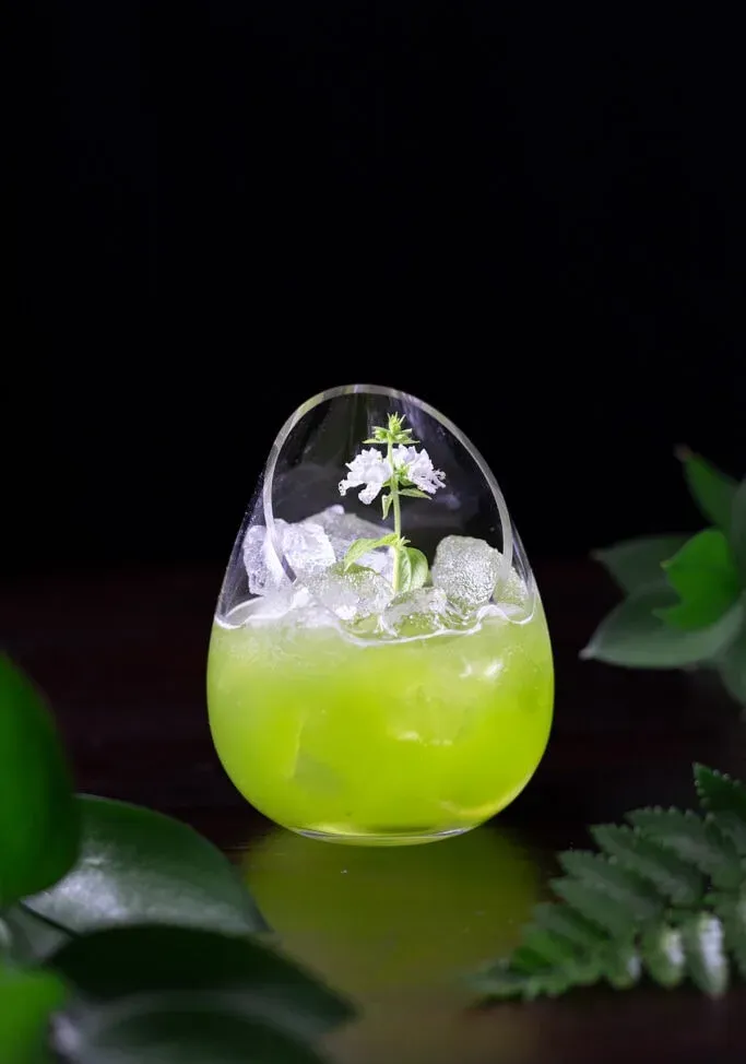 משקה ירוק בכוס מפוארת ללא גבעולים עם פרח לבן.' title=