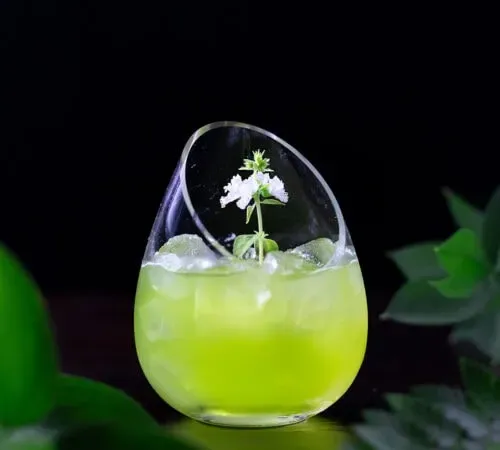 un cocktail vert dans un verre fantaisie sans pied avec une fleur blanche.' title=