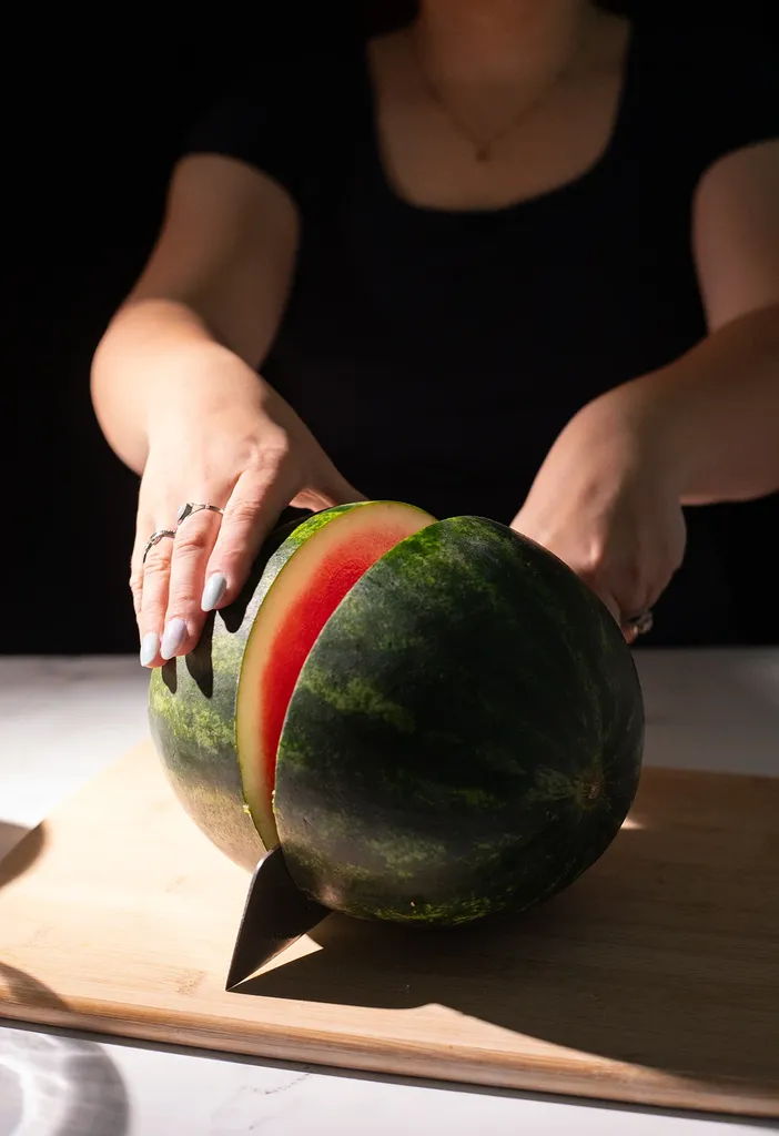 ręce kobiety przecinają okrągły arbuz na pół dużym nożem.' loading='lazy' title=