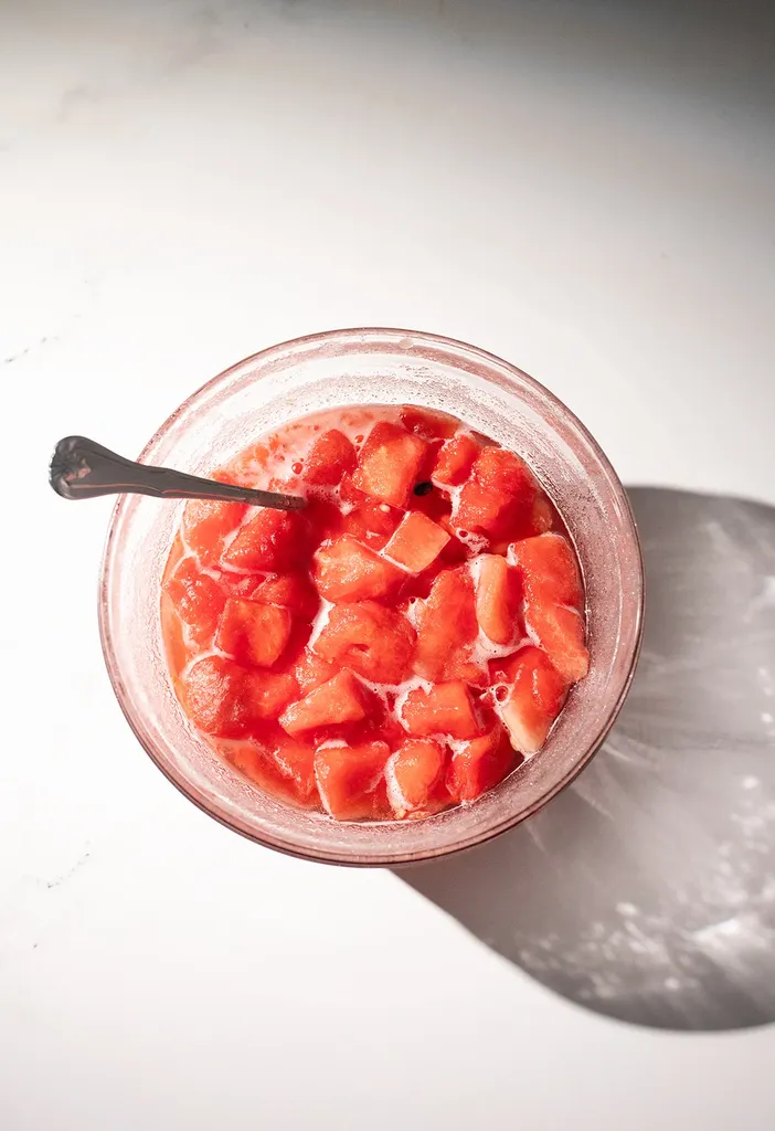 szklana butelka jasnego czerwono-różowego płynu obok plasterków arbuza i pojemnika na cukier.' title=