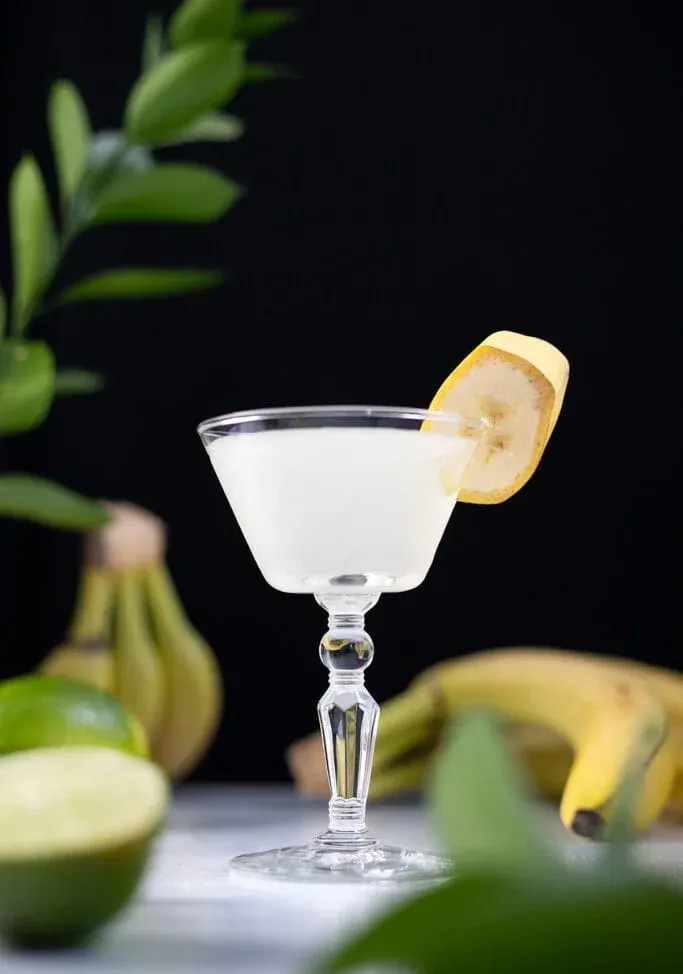 un cocktail bianco con una fetta di banana accanto a banane e lime.' title=