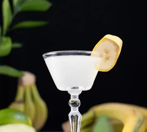 un cocktail bianco con una fetta di banana accanto a banane e lime.' title=