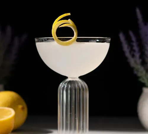 verre coupé fantaisie rempli de liquide pâle et garni de zeste de citron sur le bord.' title=