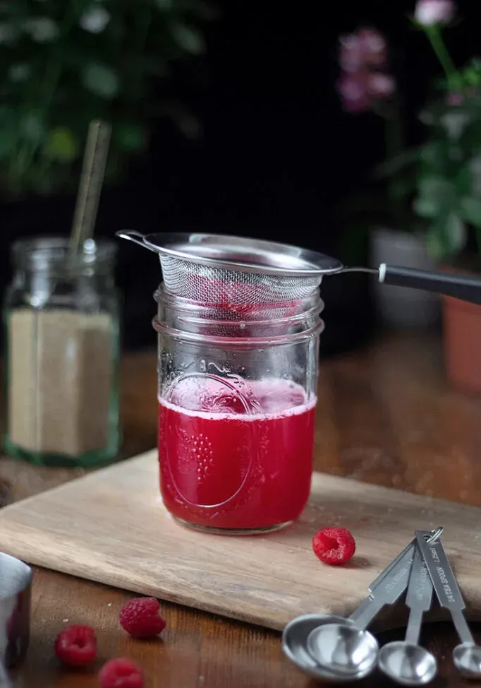 coar o xarope de framboesa através de uma peneira de malha fina em um frasco de vidro' title=