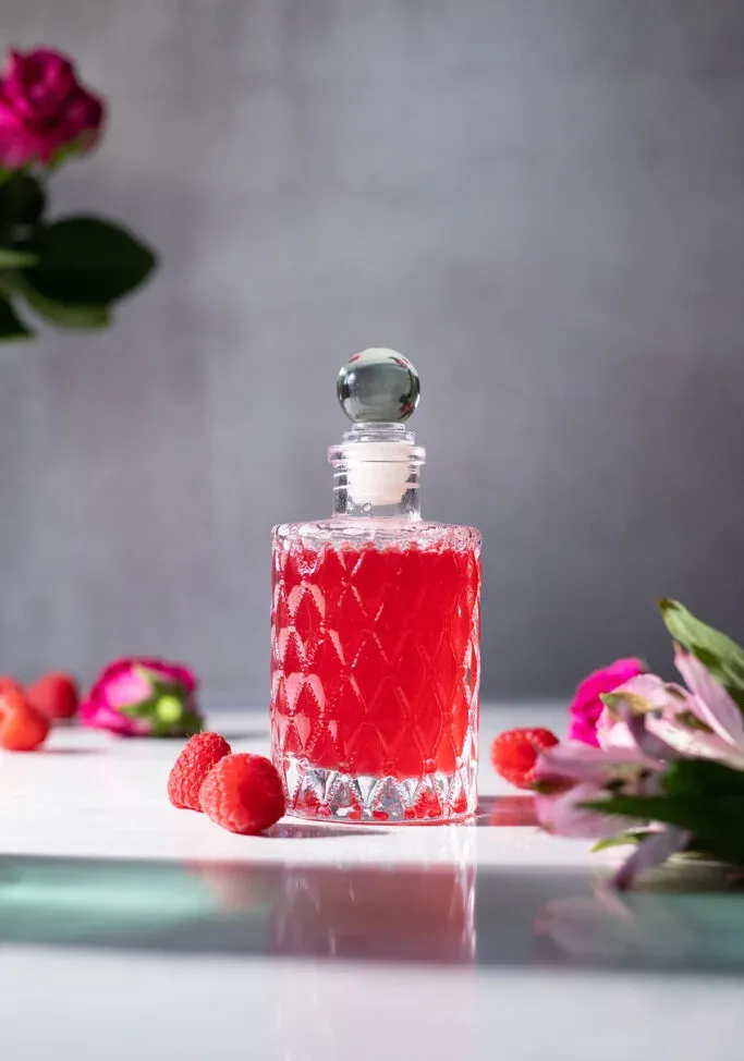 uma elegante garrafa de vidro com xarope de framboesa vermelha ao lado de frutas e flores.' loading='lazy' title=