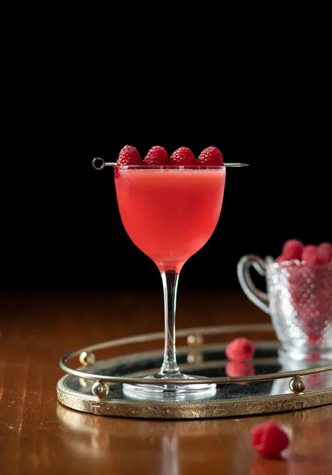 uma bebida vermelha em um copo de coquetel com framboesas em uma palheta' title=