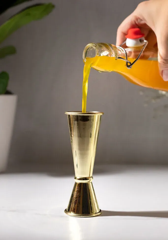 Nahaufnahme des Eingießens von Kurkuma-Sirup in einen Goldbarren-Jigger.' loading='lazy' title=