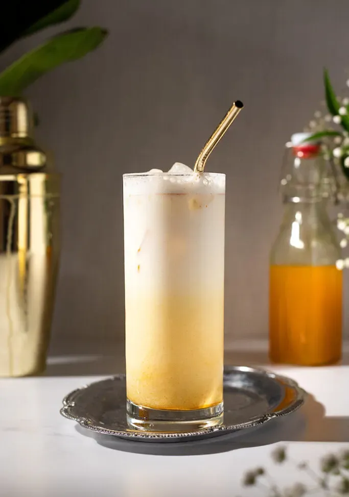 ein Glas eisgekühlte goldene Milch neben einem goldenen Shaker und einer Flasche Kurkumasirup.' title=