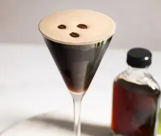 brązowy koktajl w szklance martini ozdobionej trzema ziarnami kawy.' title=