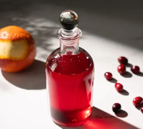 malá sklenená fľaša červeného sirupu vedľa brusníc a pomaranča.' title=