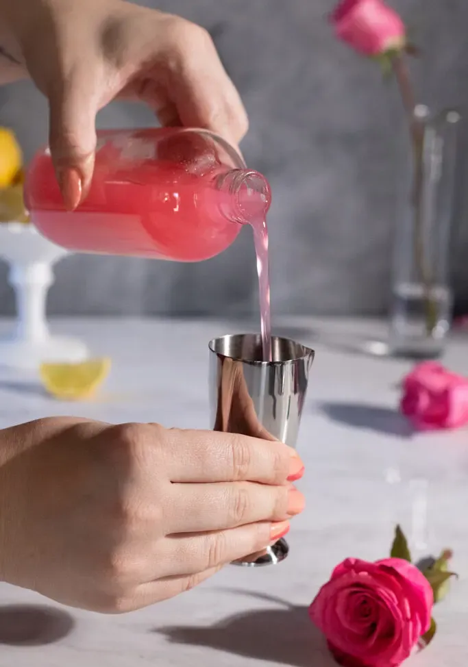 nalievanie ružového sirupu z fľaše do striebornej tyčinky vedľa ruží.' title=