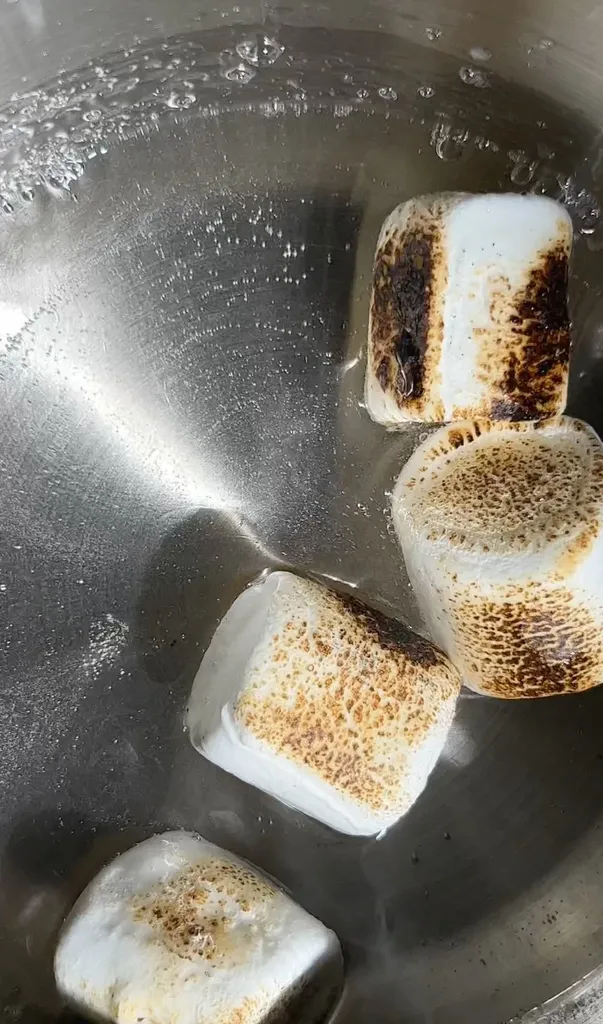 quattro marshmallow tostati galleggianti in un liquido trasparente in una casseruola di metallo.' loading='lazy' title=
