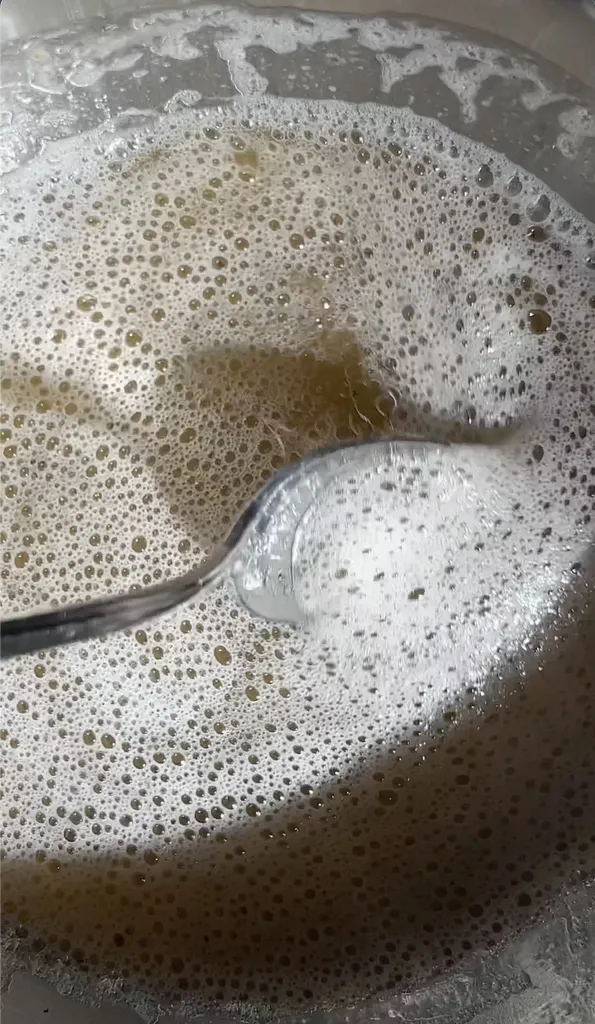 cucchiaio di metallo immerso in un liquido color oro con schiuma bianca sulla parte superiore.' loading='lazy' title=