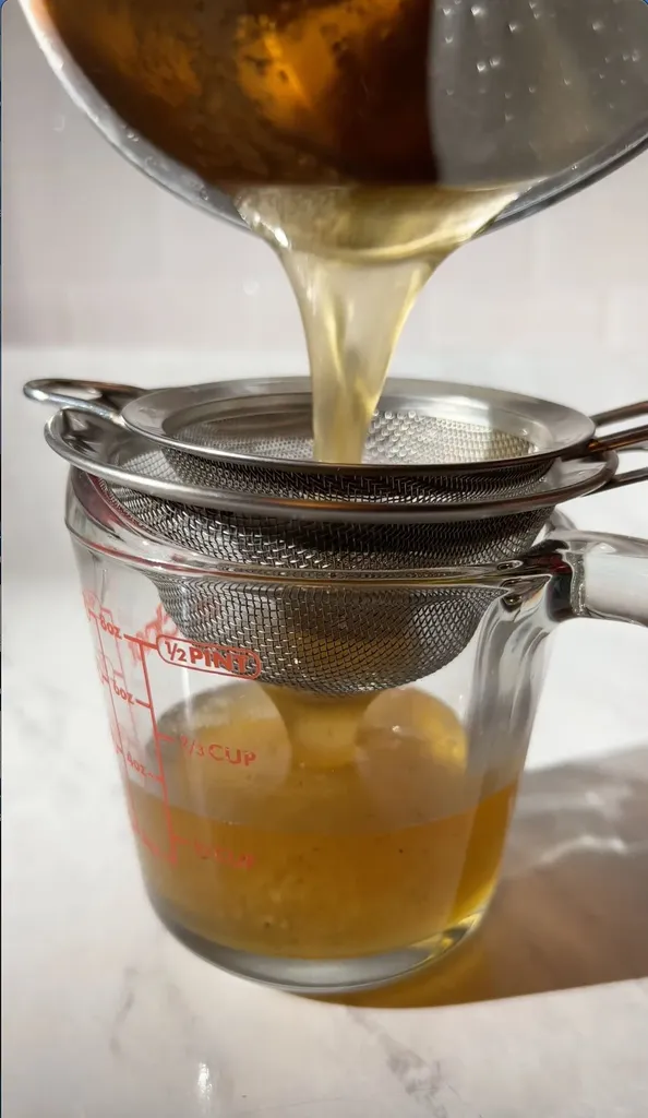versando lo sciroppo dorato attraverso un colino a maglia fine in un misurino di vetro.' title=
