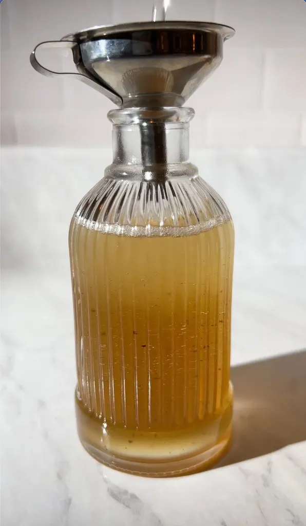 versando lo sciroppo marrone dorato attraverso un imbuto in una piccola bottiglia di vetro.' title=