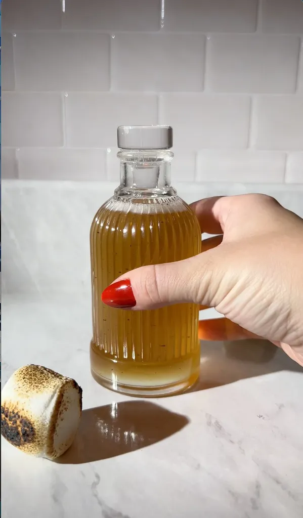 la mano di una donna che tiene una bottiglia di sciroppo dorato accanto a un marshmallow tostato.' title=