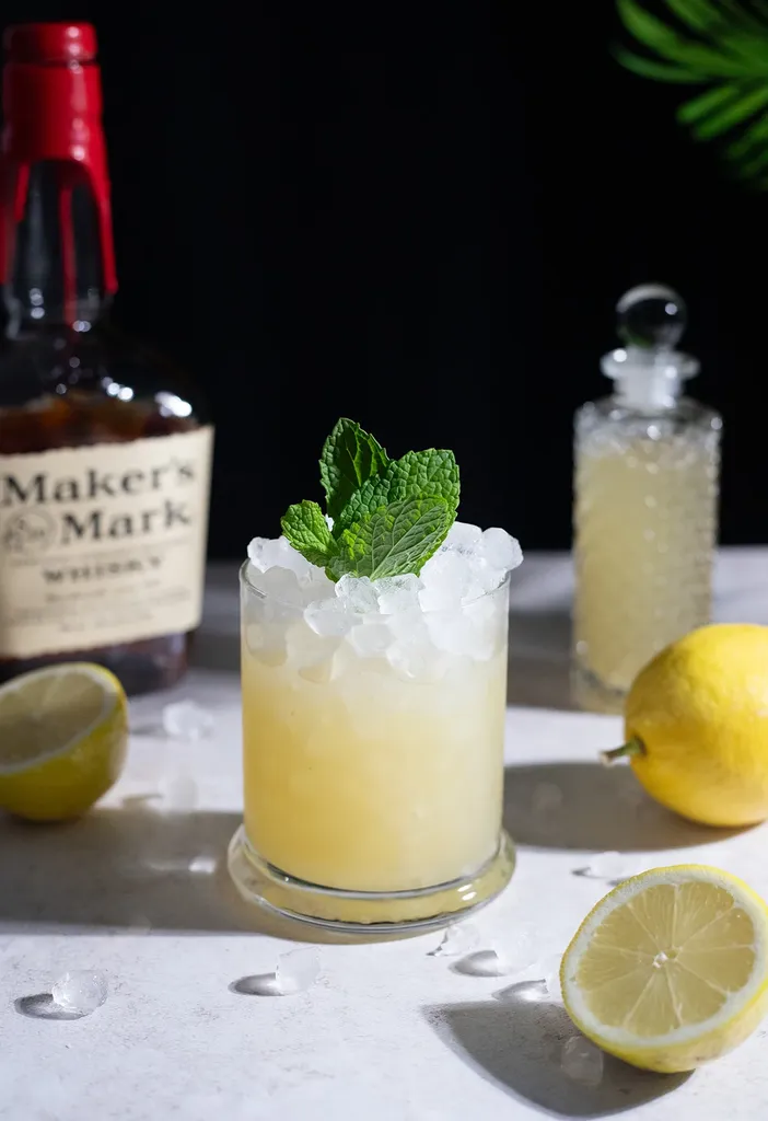Cóctel de color amarillo pálido en un vaso bajo adornado con menta junto a una botella de Maker's Mark.' loading='lazy' title=