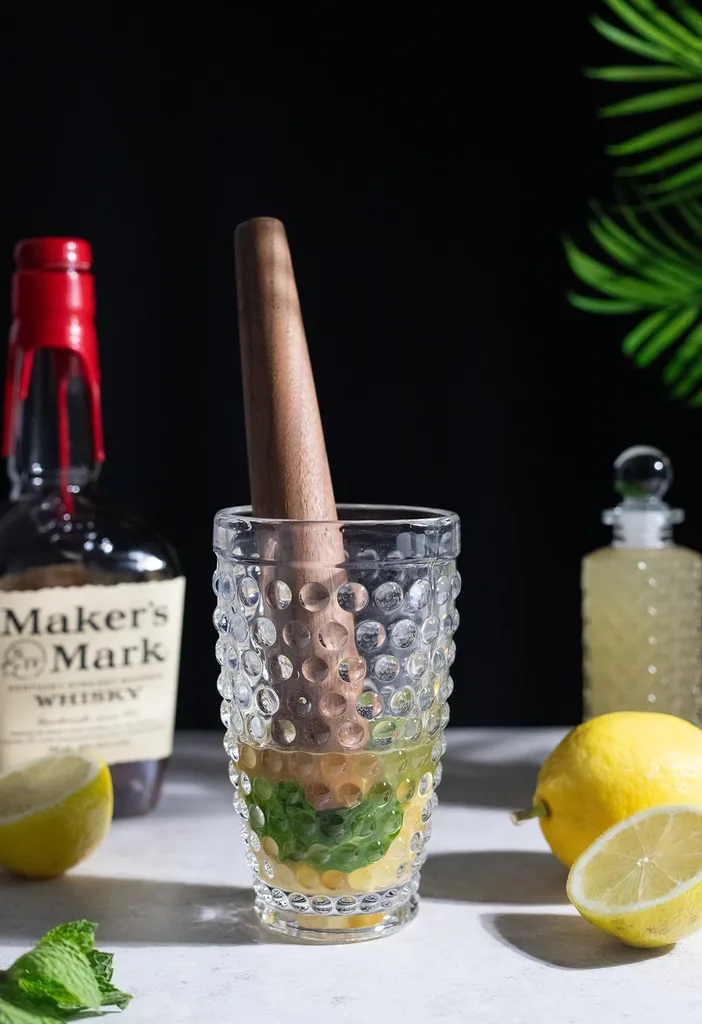 Muddler de madera colocado en una coctelera de vidrio llena de líquido amarillo y hojas de menta.' title=